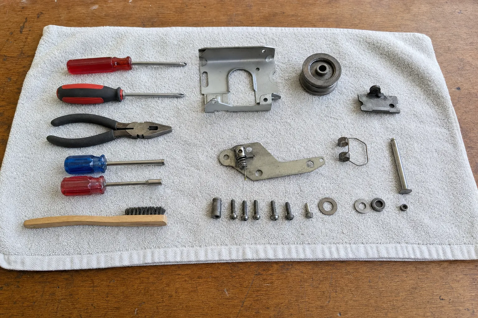 Screwdrivers, pliers and a wire brush beside a stamped bracket, a pulley wheel, springs, a pin and washers on a towel