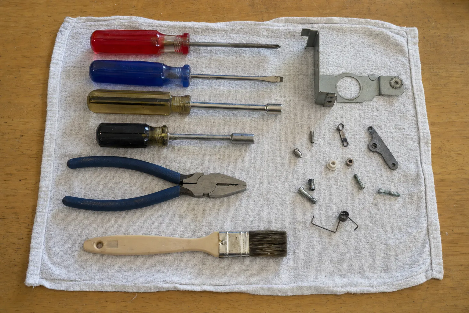Four drivers, blue-handled pliers and a paintbrush beside a bracket with a round cut-out, springs and small spacers
