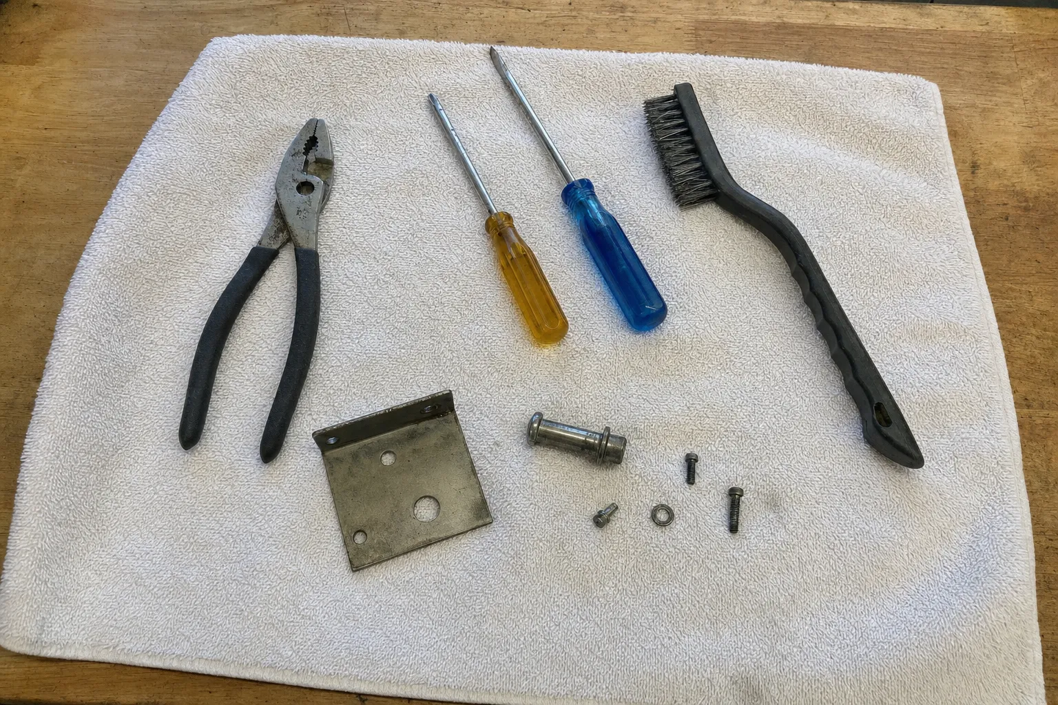Slip-joint pliers, two screwdrivers, a plastic-handled wire brush, a flat steel bracket and a shouldered pin on a white towel